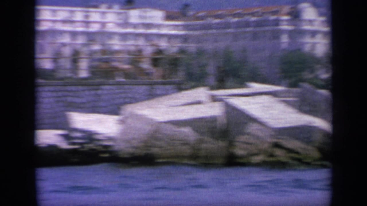 1963 Greece 8mm frame showing blue harbor water, a stone breakwater, and pale waterfront buildings beyond the shore.