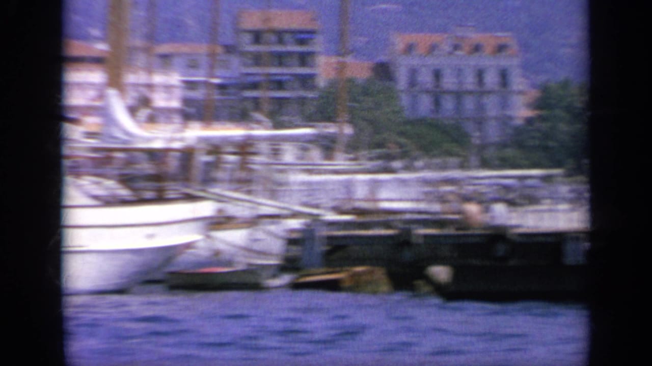 1963 Greece harbor view with a moored white boat, masts, quay structures, and waterfront buildings in the background.