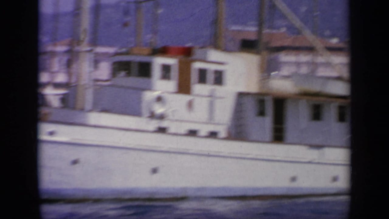 Close 8mm view of a large white yacht in a Greek harbor, with cabin windows, railings, masts, and waterfront structures behind it.