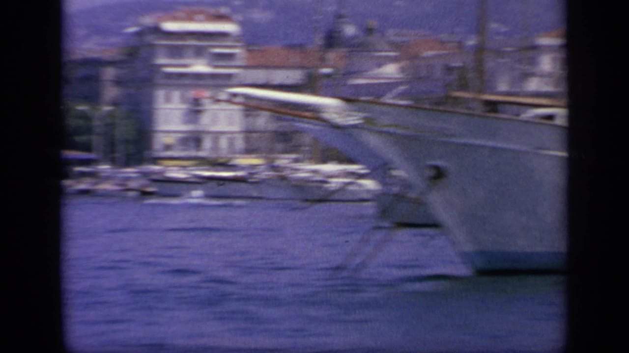 1963 Greece frame showing the bow of a large white yacht on blue harbor water, with shoreline buildings and other boats in the distance.