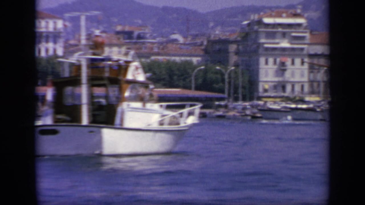 White motorboat on blue harbor water with waterfront buildings and hills behind it.