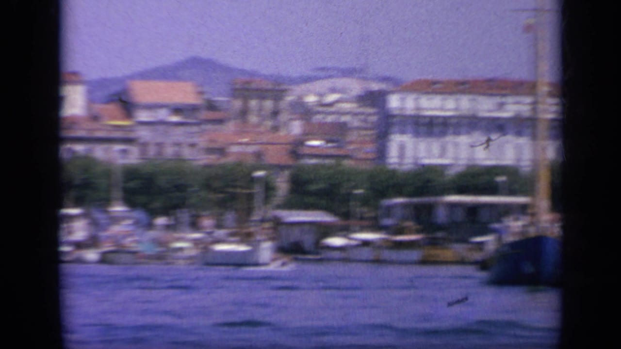 1963 Greece waterfront seen across blue harbor water, with small boats along the quay, urban buildings, and hills rising behind them.