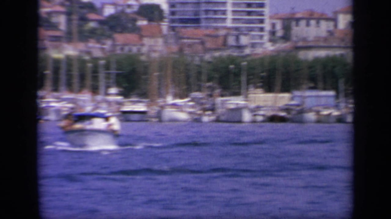 1963 Greece harbor frame with a small boat on blue water, marina masts, waterfront structures, and hillside buildings in the distance.