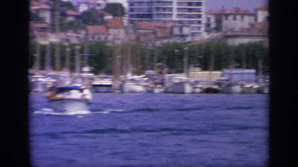 1963 Greece harbor frame with a small boat on blue water, marina masts, waterfront structures, and hillside buildings in the distance.