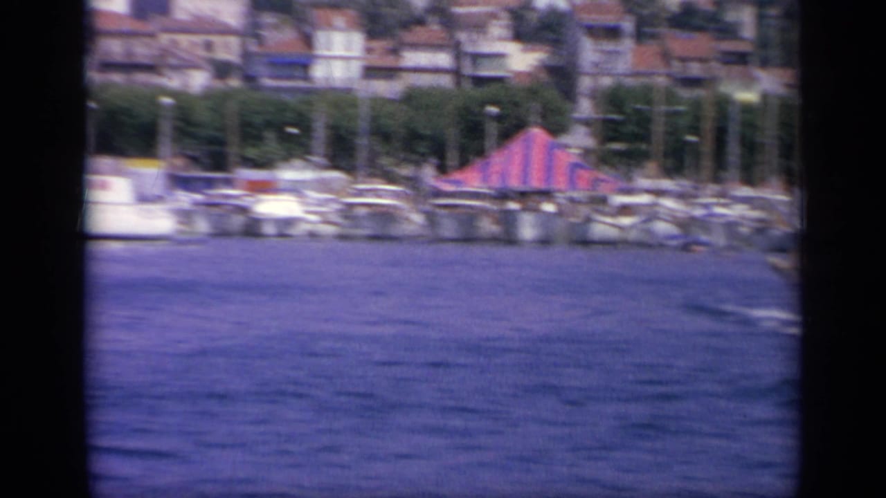 1963 Greece harbor view showing moored boats, a red-and-blue striped tent near the quay, waterfront trees, buildings, and distant hills.