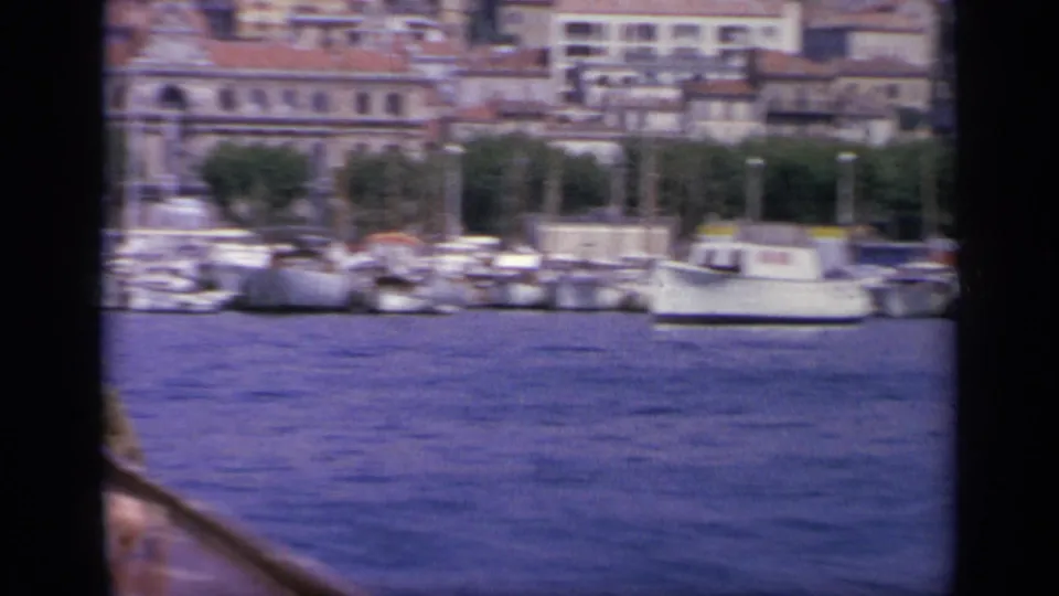 1963 Greece 8mm view of blue harbor water, moored boats, and waterfront buildings seen past dark boat or window framing.