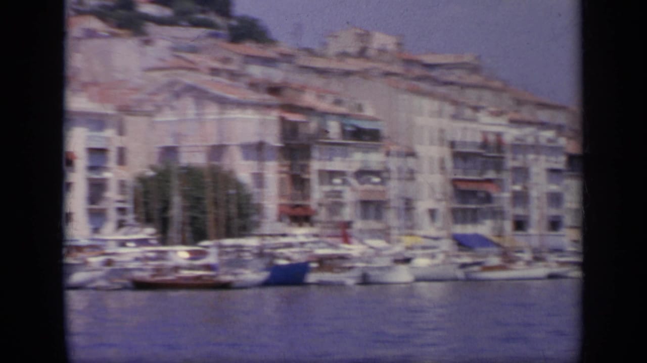 1963 Greece 8mm frame showing moored boats, a harbor quay, and pale waterfront buildings with red-tiled roofs.