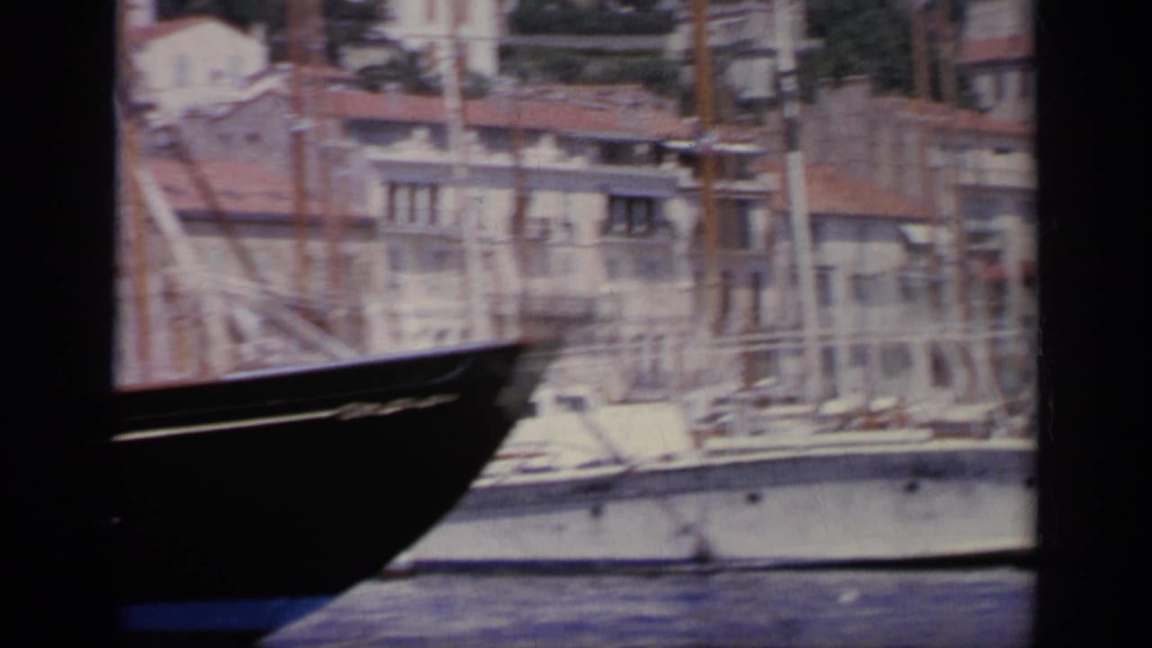 1963 Greece harbor frame with a white boat, masts, quay buildings, and dark foreground boat or window edges.