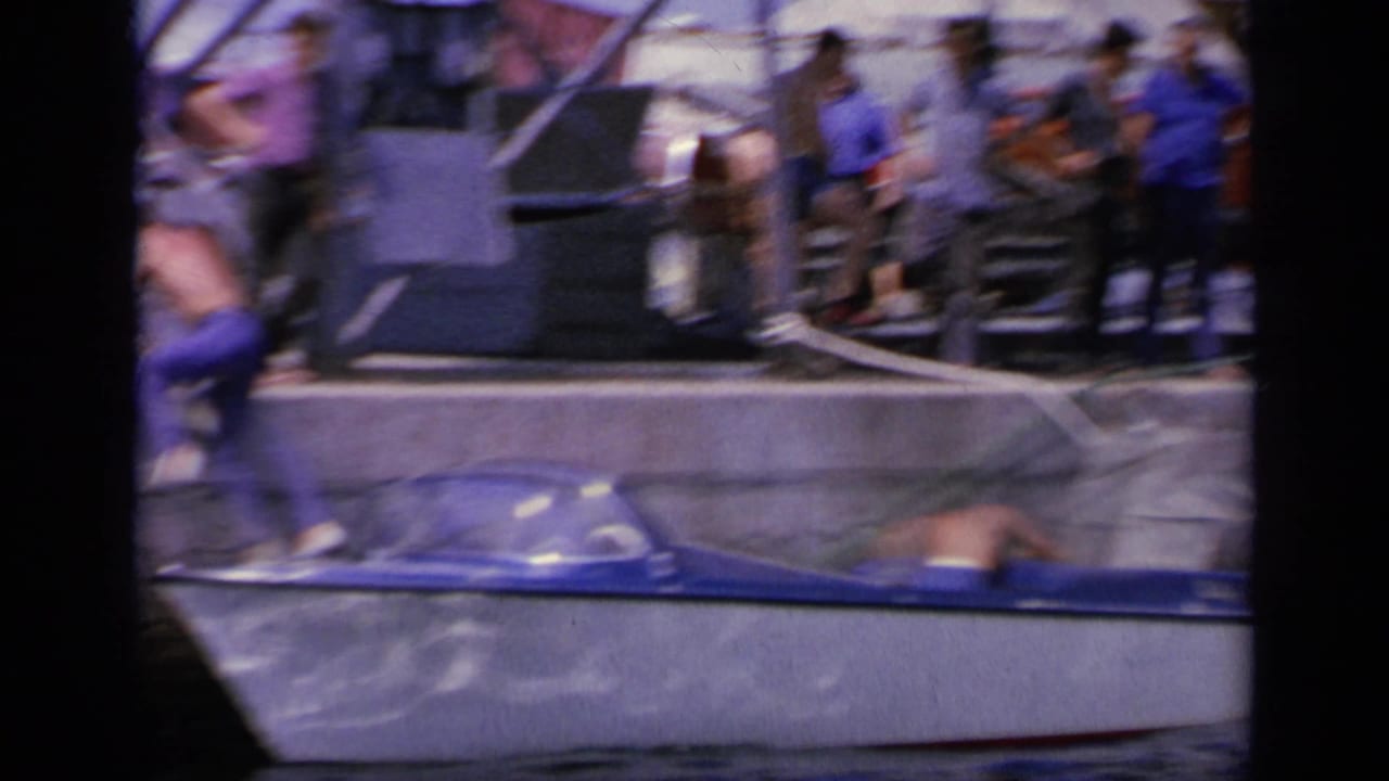 1963 Greece waterfront frame showing people on the quay near a small blue boat, with legs and faces partly visible.
