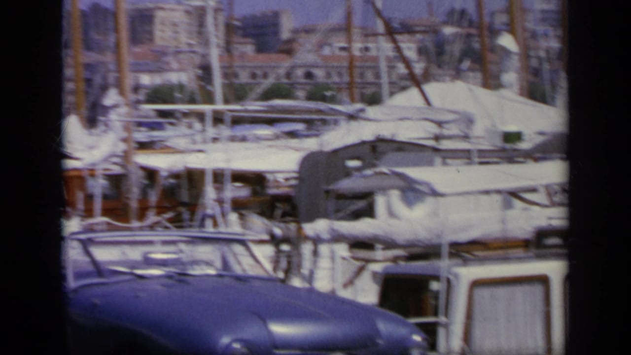 Moored sailboats and a blue car along a crowded harbor waterfront.