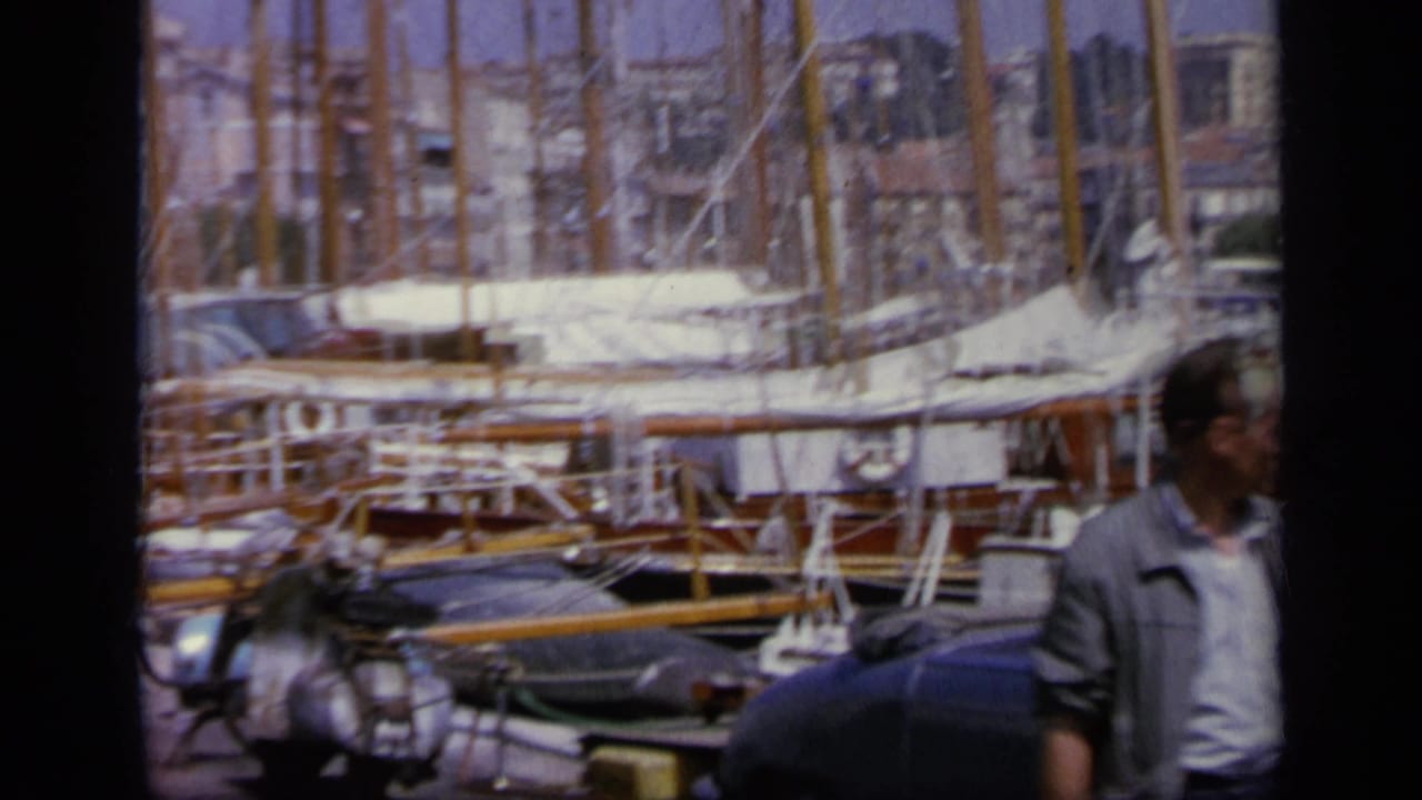 1963 Greece harbor view with a man at the edge of frame, moored sailboats, a scooter, and sunlit buildings in the background.