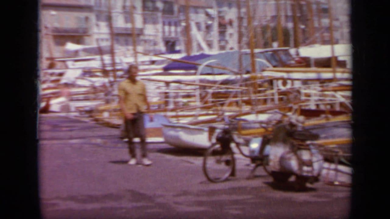 1963 Greece waterfront frame with a person standing near parked scooters, moored sailboats, and a dense marina backdrop.