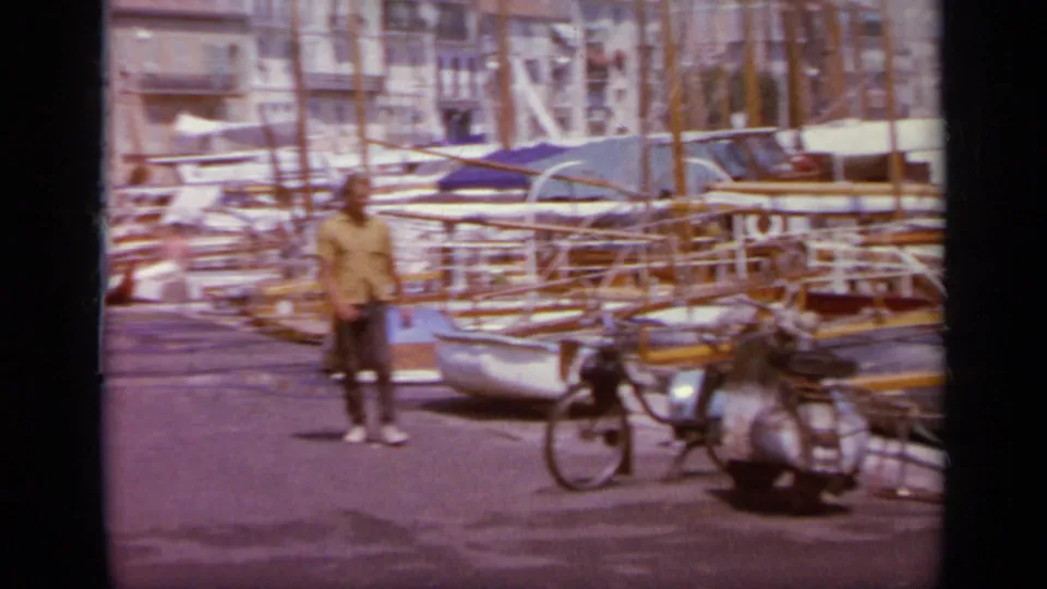 1963 Greece waterfront frame with a person standing near parked scooters, moored sailboats, and a dense marina backdrop.