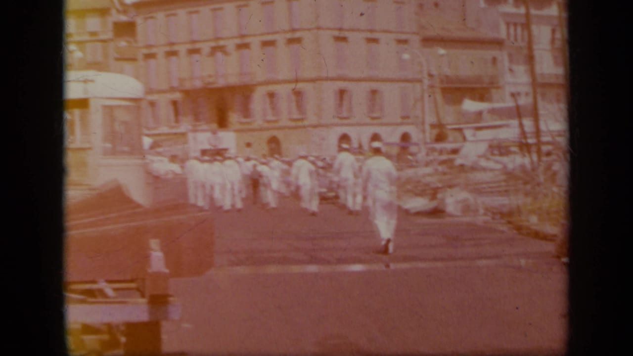 1963 Greece harbor frame showing sailors in white uniforms walking near a large waterfront building and moored boats.