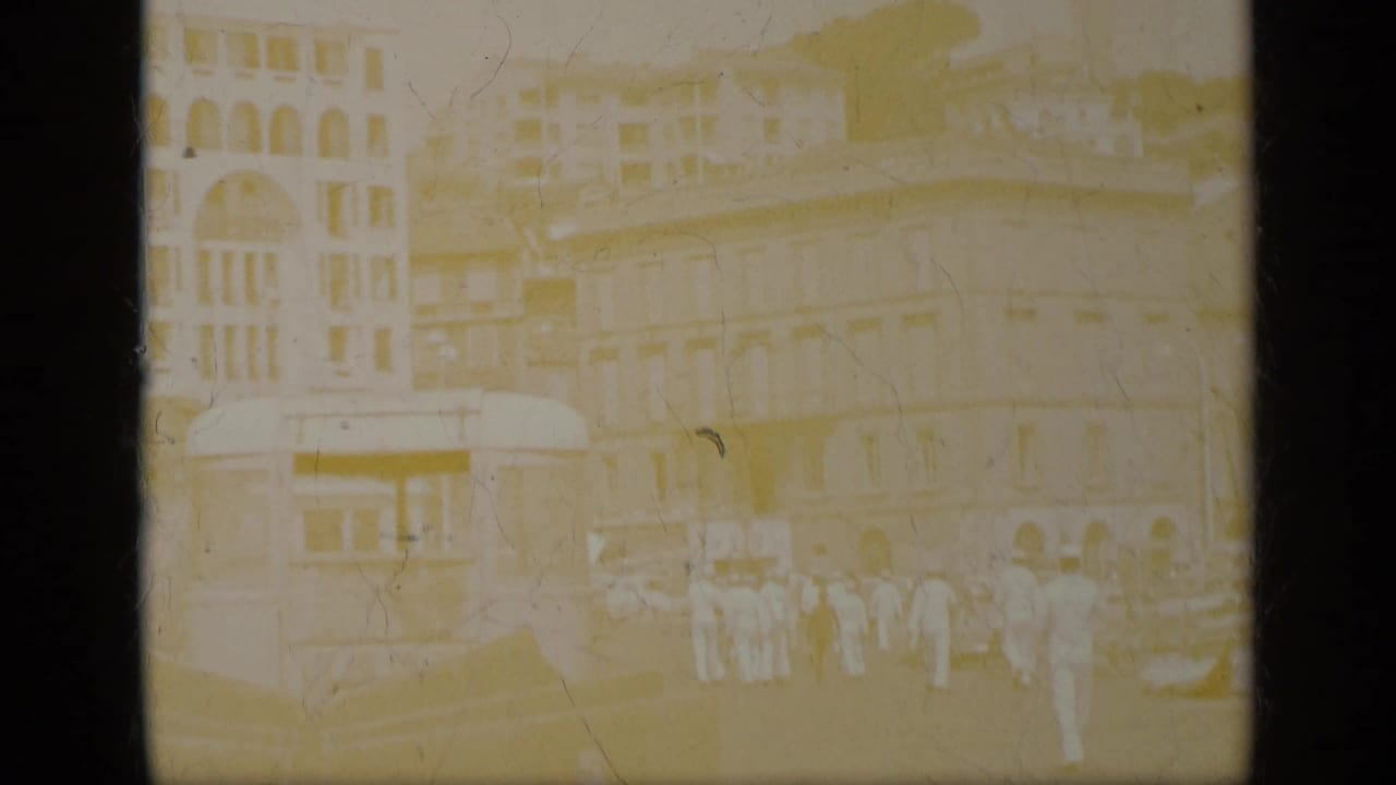 1963 Greece home-movie frame showing men in white uniforms near a tram and multi-story street facades, with heavy fading and soft focus.