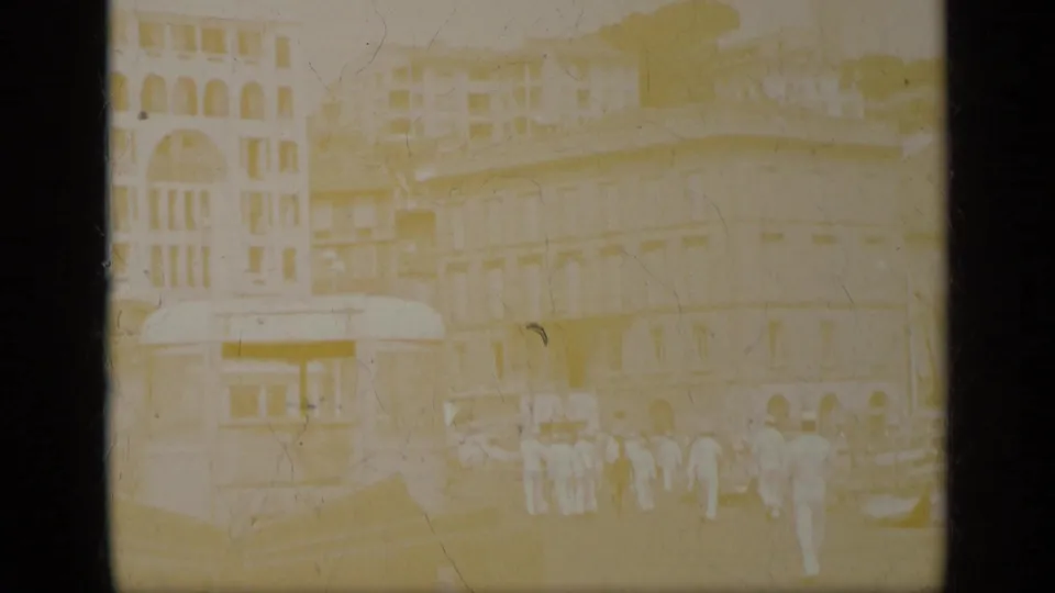 1963 Greece home-movie frame showing men in white uniforms near a tram and multi-story street facades, with heavy fading and soft focus.