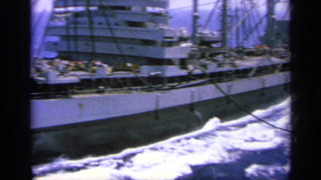 The camera continues along the vessel's side, showing midship structures, railings, rigging, and white water breaking beside the hull.