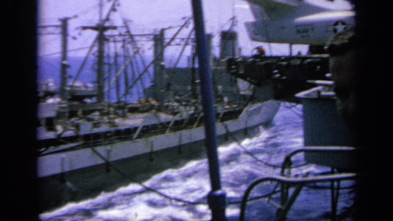 1963 Greece home-movie view of a gray ship and wake beside carrier deck equipment and a parked jet edge.