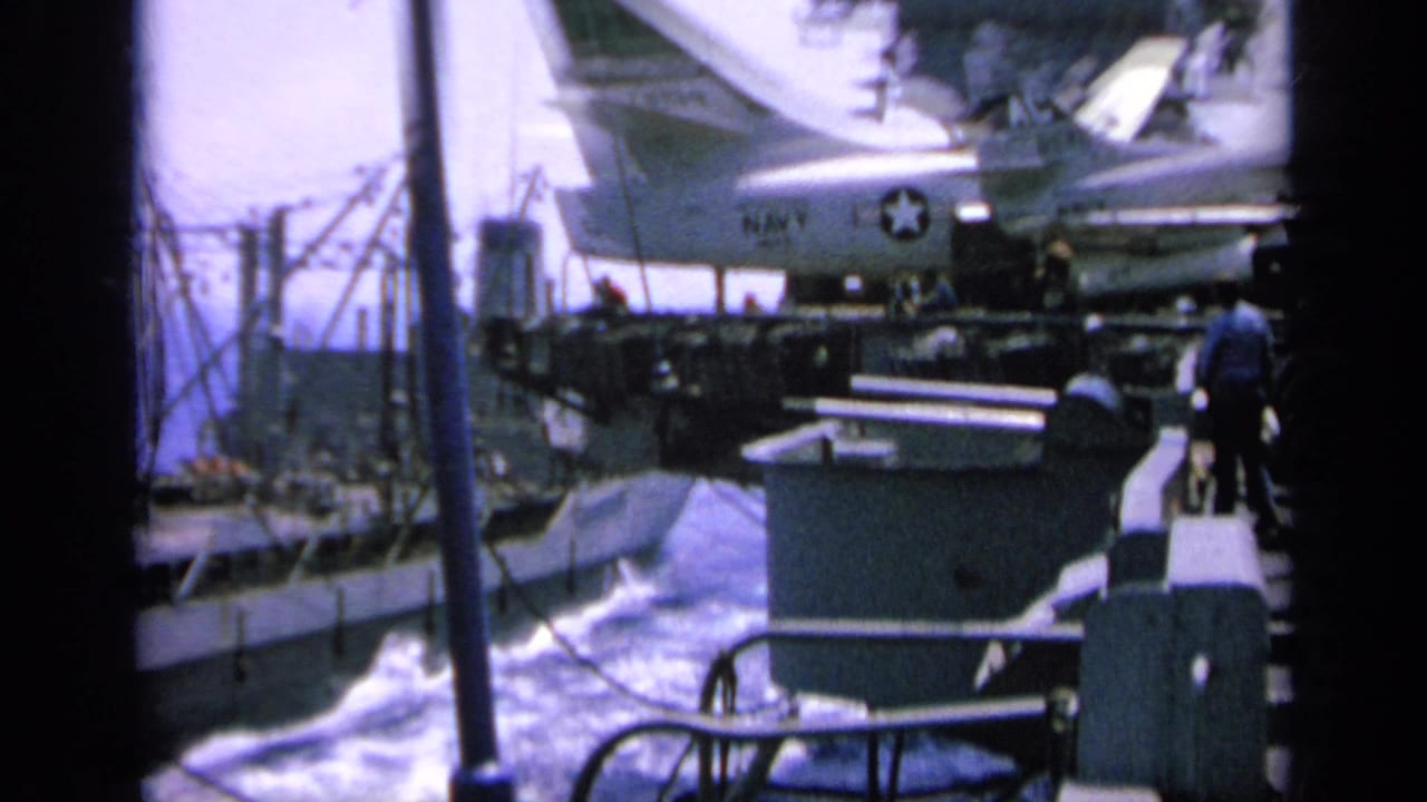 1963 Greece frame showing a carrier deck, parked jet with U.S. star insignia, small people, and a ship passing alongside.