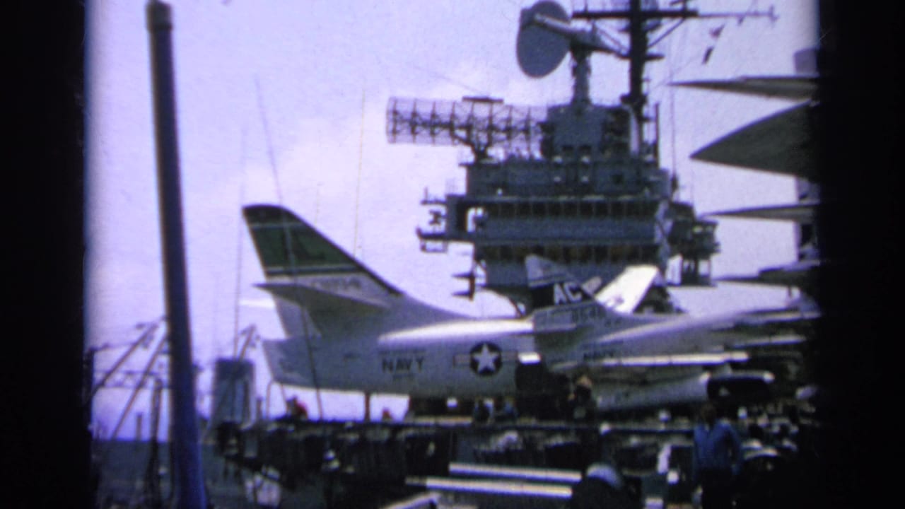 1963 Greece-labeled home-movie frame showing a Navy jet parked on a carrier deck below the ship island and radar equipment.
