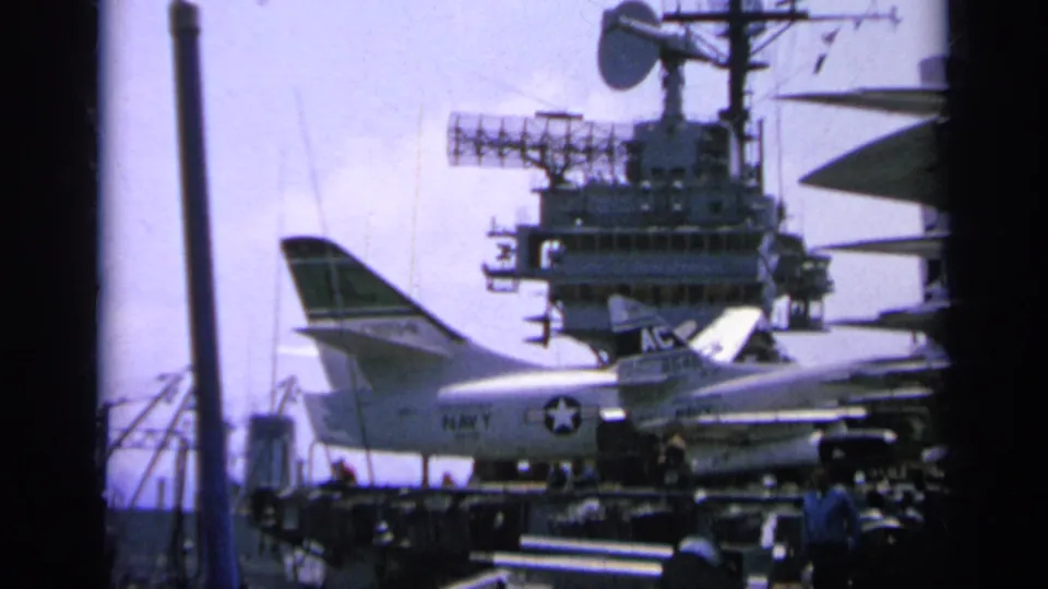 1963 Greece-labeled home-movie frame showing a Navy jet parked on a carrier deck below the ship island and radar equipment.
