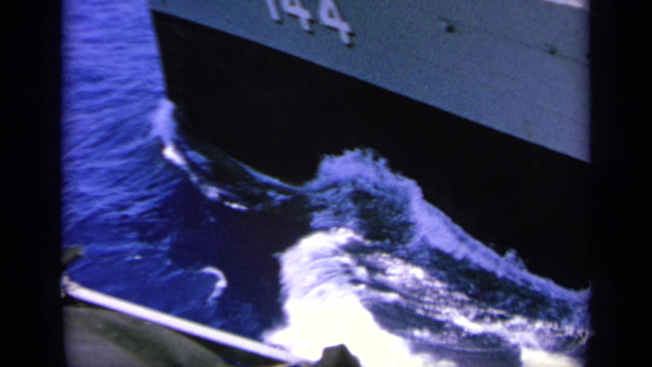 The ship hull marked 144 fills the frame as white spray churns against deep blue water during the naval passage.