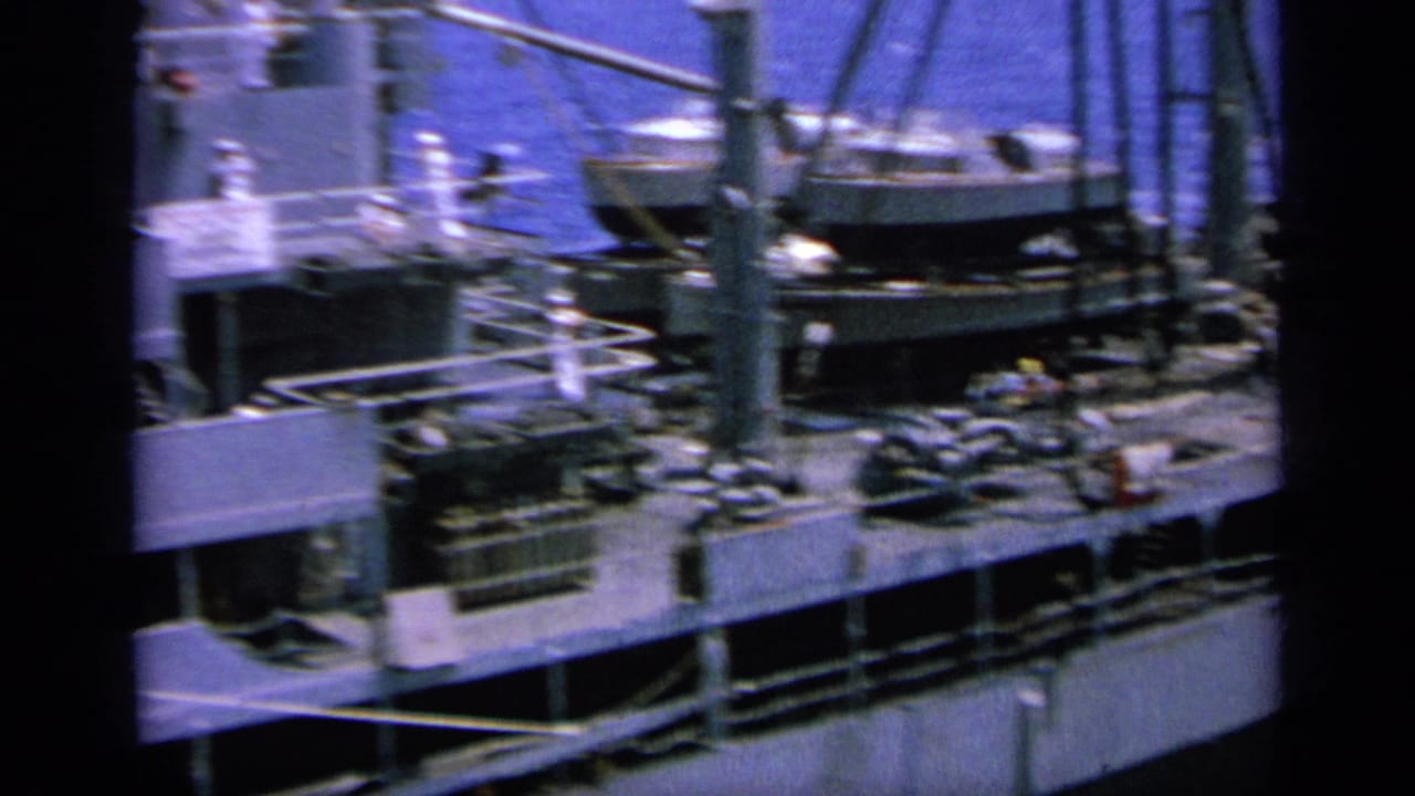 Large pale ship deck seen from a nearby vessel, with rigging and equipment under bright sun.