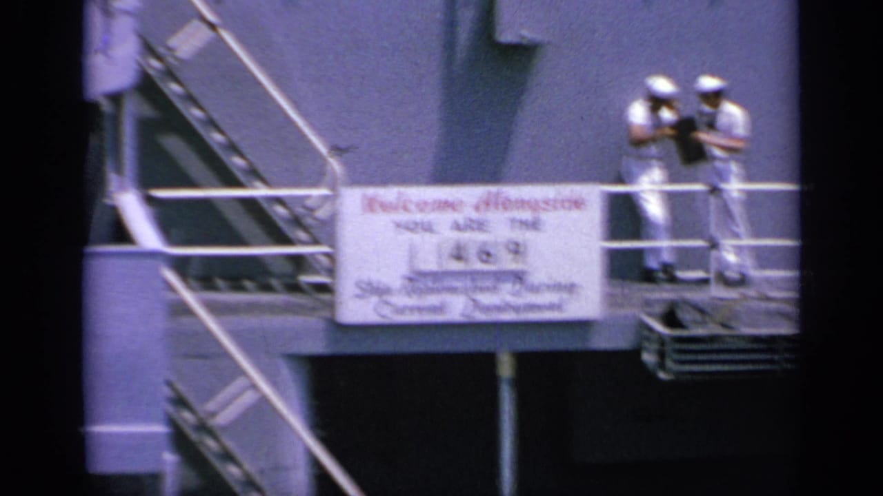 1963 Greece 8mm frame showing two sailors paused on deck near railings, stairs, and the replenishment sign mounted below them.