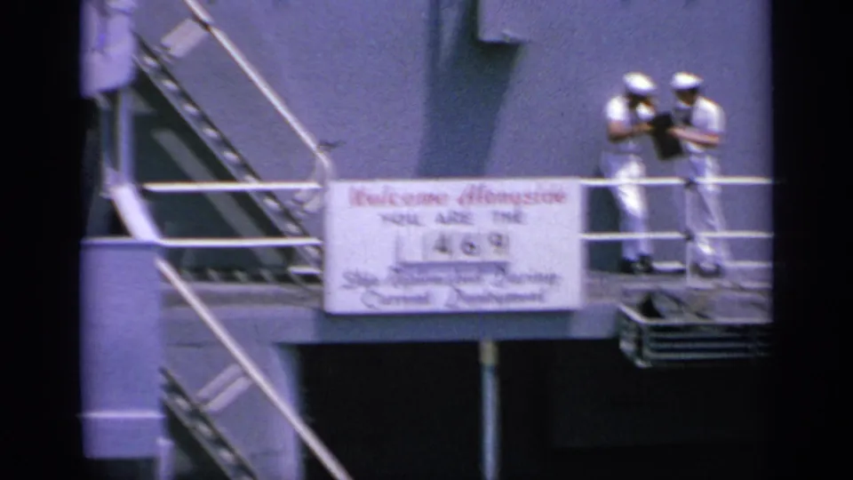 1963 Greece 8mm frame showing two sailors paused on deck near railings, stairs, and the replenishment sign mounted below them.