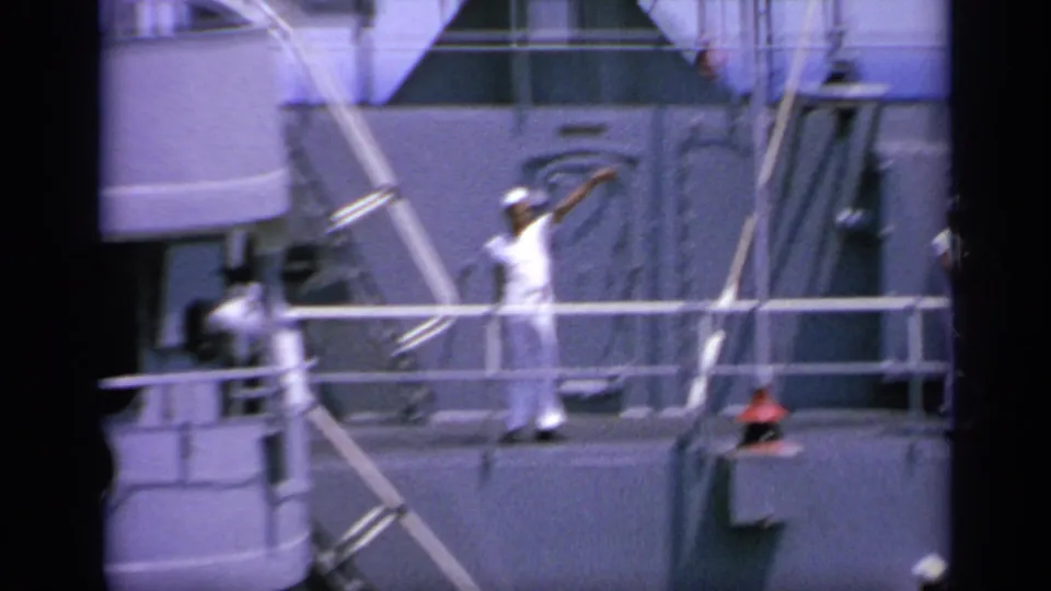 1963 Greece 8mm frame showing a sailor waving on a naval ship deck, with railings, rigging or cables, and another crew member nearby.