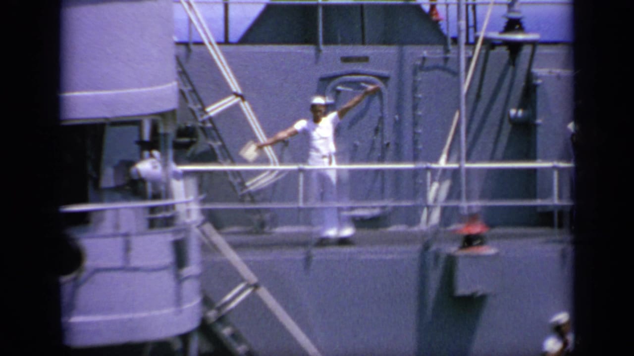 Sailor in white uniform raises signal flags on a gray ship deck.