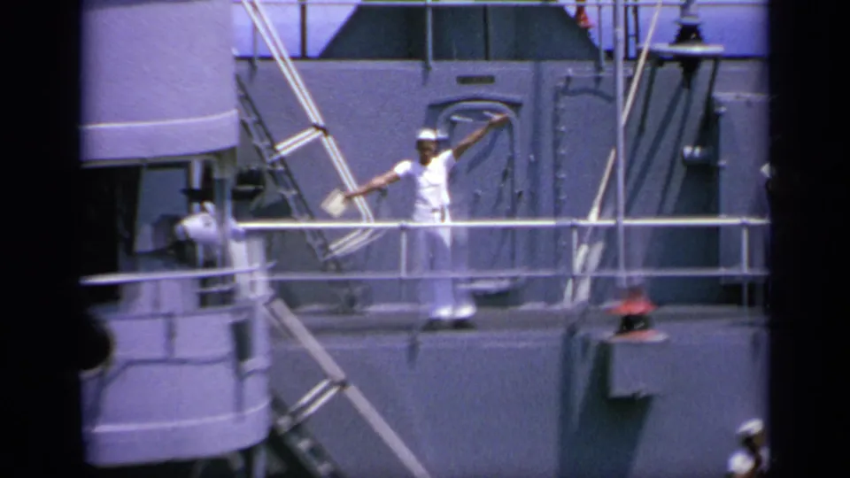 In 1963 Greece footage, a sailor in white uniform stands by railings on a naval ship and raises signal flags.