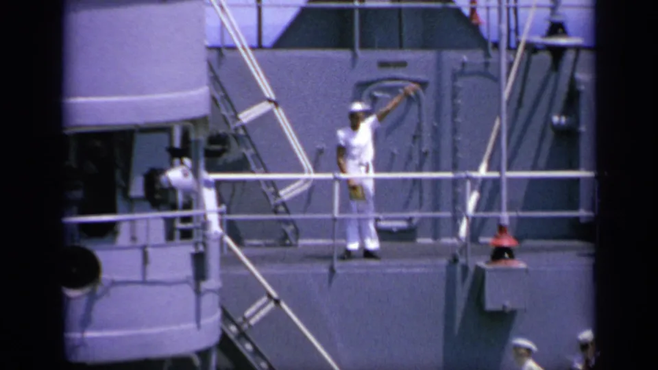 A white-uniformed sailor on a naval ship in Greece continues a flag signal near railings and gray superstructure.