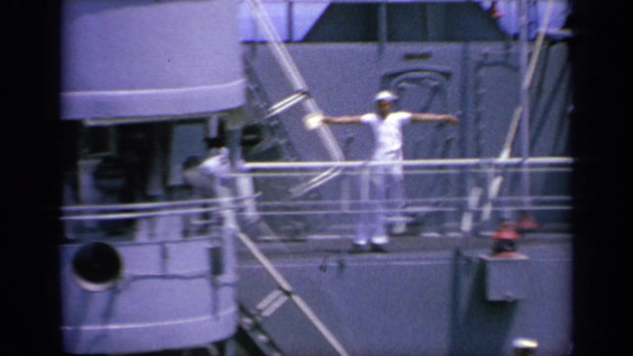 The sailor appears with arms extended in a flag-signal position on the gray naval ship deck in 1963 Greece footage.