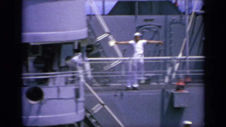 The sailor appears with arms extended in a flag-signal position on the gray naval ship deck in 1963 Greece footage.