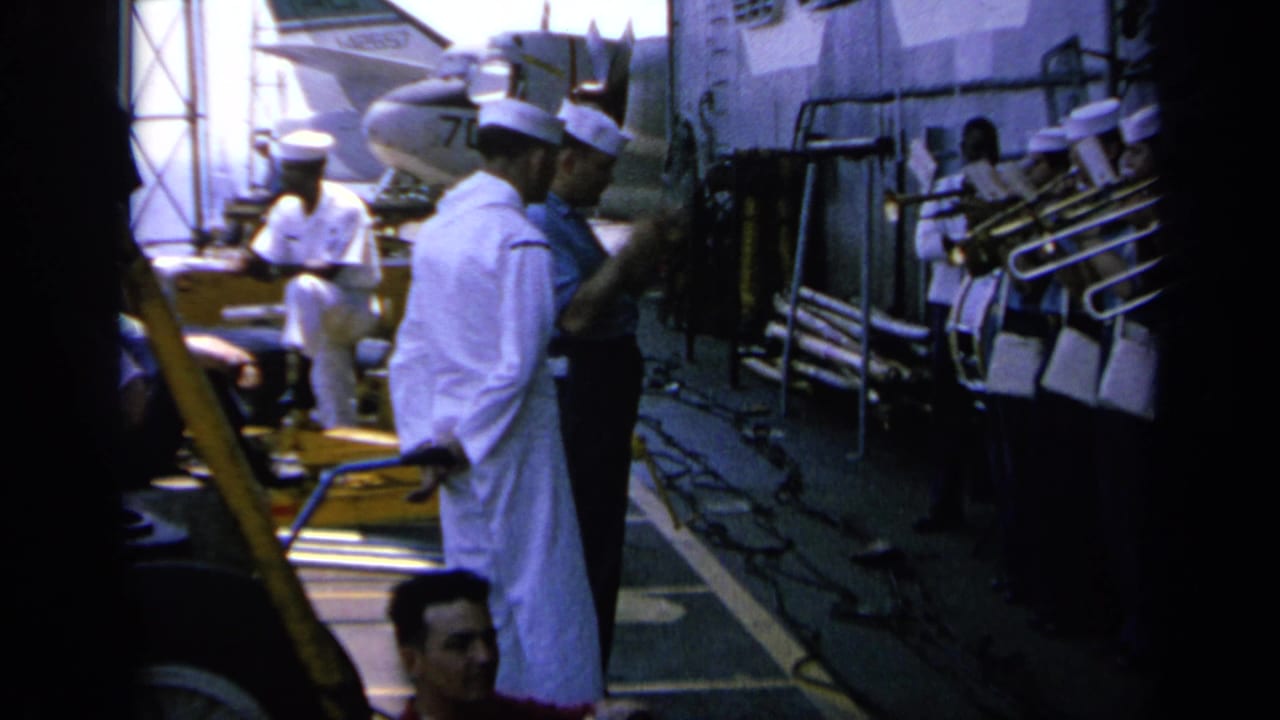 Deck-level view of uniformed crew members on a naval ship in Greece, with railings, pipes, and equipment visible nearby.
