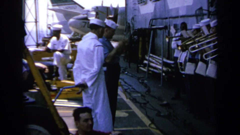 Deck-level view of uniformed crew members on a naval ship in Greece, with railings, pipes, and equipment visible nearby.