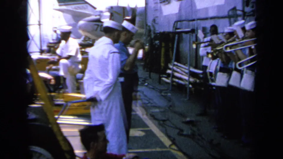 1963 Greece home-movie frame showing U.S. Navy band members on deck with trombones and aircraft visible behind them.