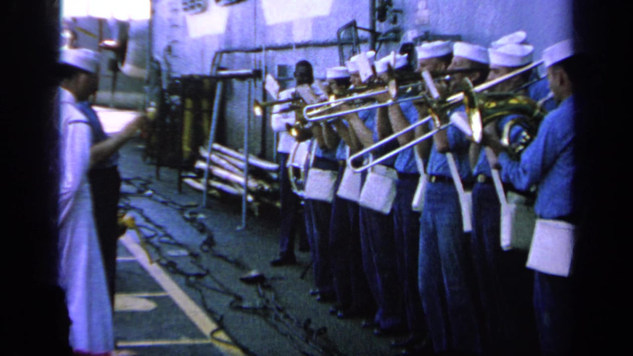 U.S. Navy musicians perform on a ship deck in Greece, with trombones, white caps, and blue shirts visible.