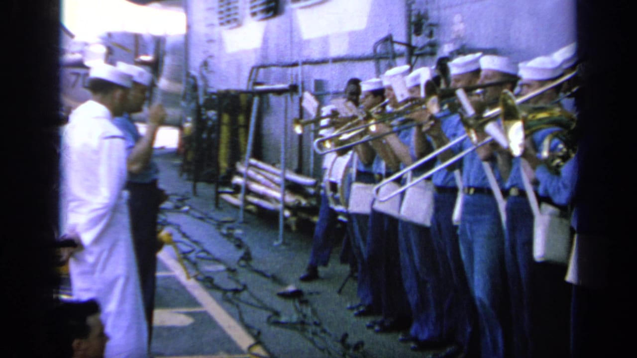 A conductor in white stands near sailors playing brass instruments on a ship deck.