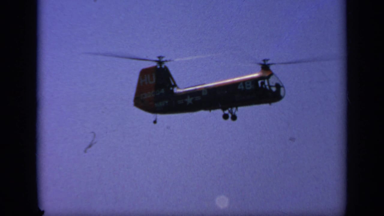 1963 Greece home-movie frame of a dark military helicopter in flight, with partial markings visible on the tail and fuselage.
