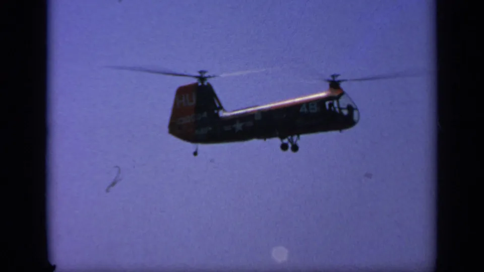 1963 Greece home-movie frame of a dark military helicopter in flight, with partial markings visible on the tail and fuselage.