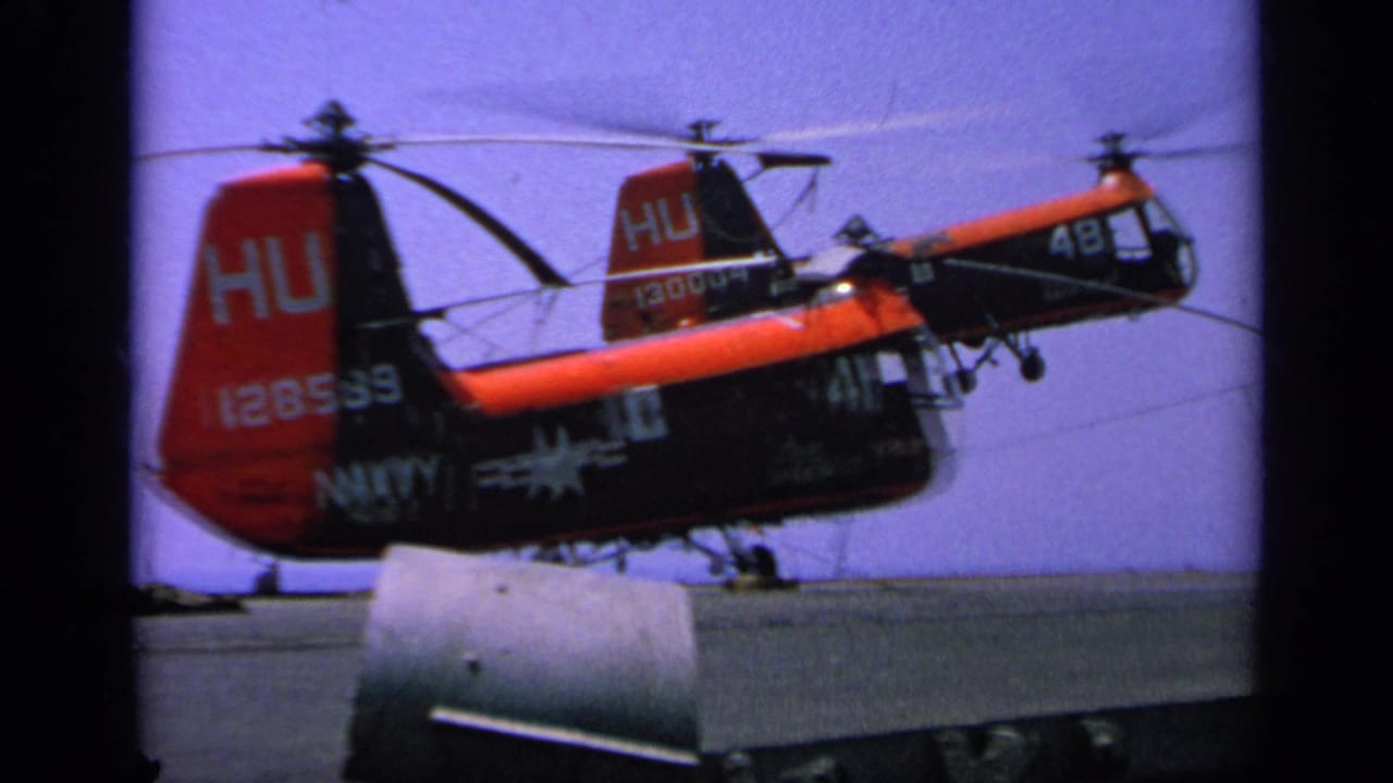 1963 Greece frame of a red and black Navy helicopter hovering low over a carrier deck, with another helicopter partly visible behind it.