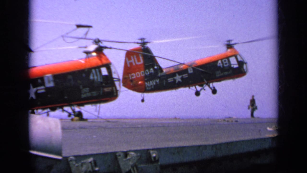 A 1963 Greece carrier-deck frame shows a red and black Navy helicopter moving above the deck as a uniformed crew member stands to the side.