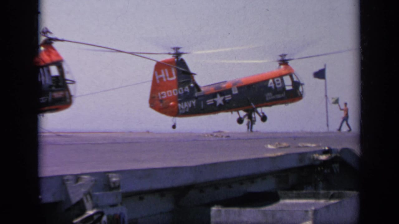 1963 Greece 8mm frame showing a red and black Navy helicopter hovering over a carrier deck, with crew figures and deck flags visible nearby.