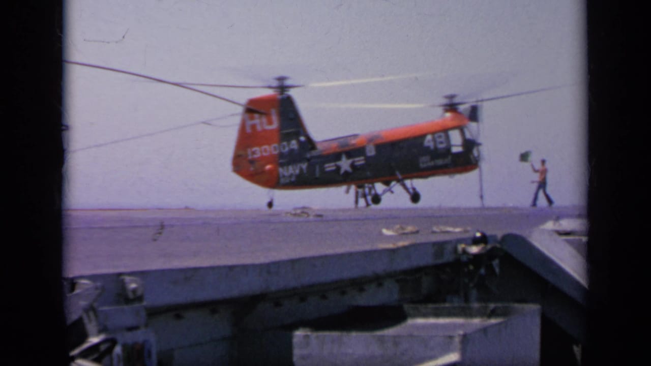 1963 Greece 8mm frame showing a red and dark Navy HU-1 helicopter descending toward a ship deck with crew guidance visible.