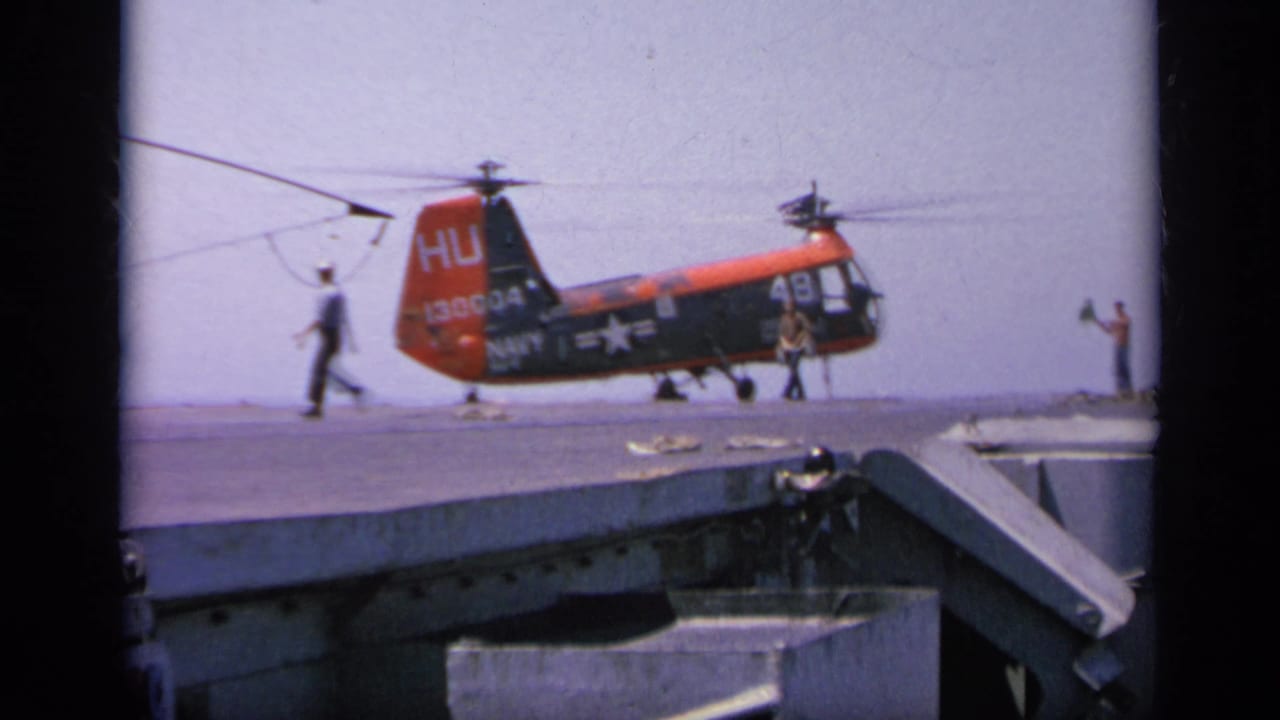1963 Greece frame of a red and dark Navy HU-1 helicopter on a ship deck, with crew nearby and rotors still blurred.