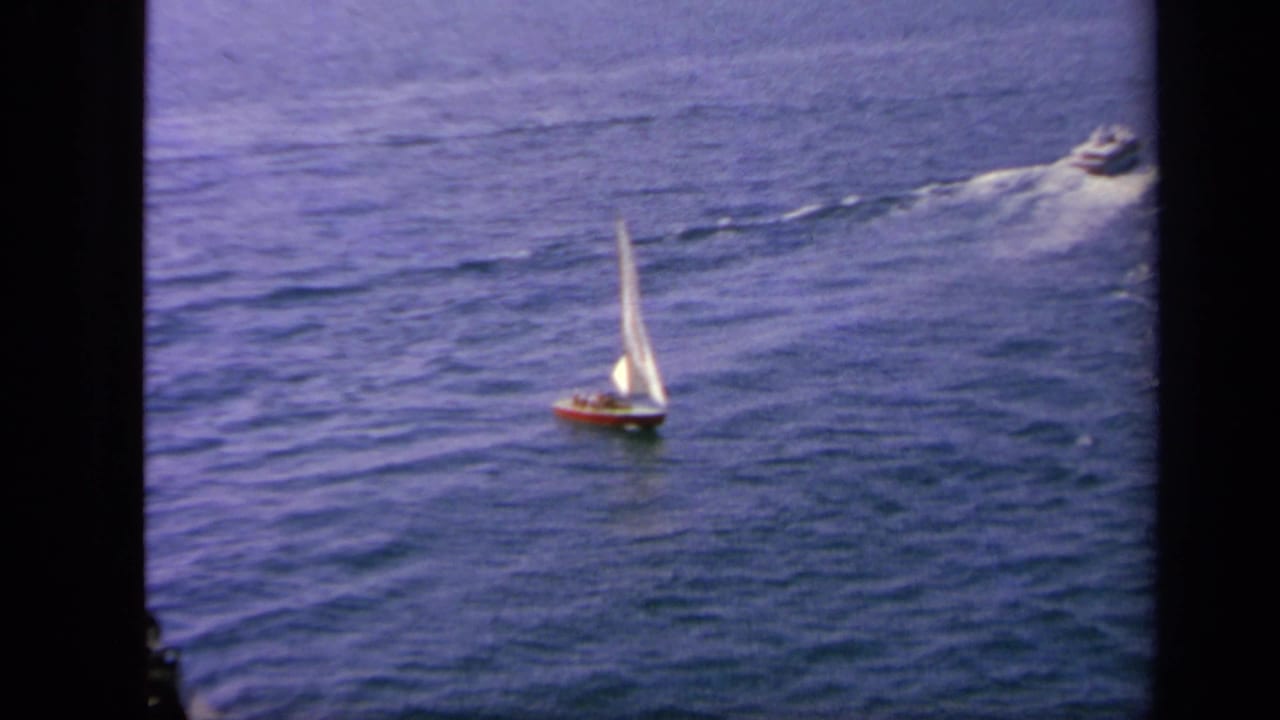 1963 Greece-area ocean view with a small sailboat in the foreground and a motorboat moving across the water behind it.