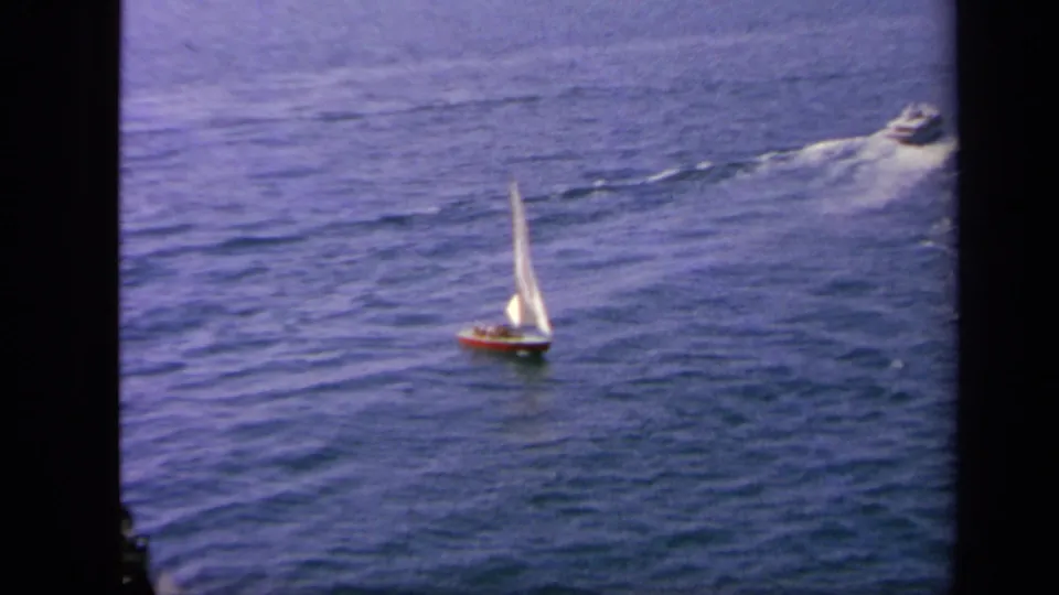 1963 Greece-area ocean view with a small sailboat in the foreground and a motorboat moving across the water behind it.
