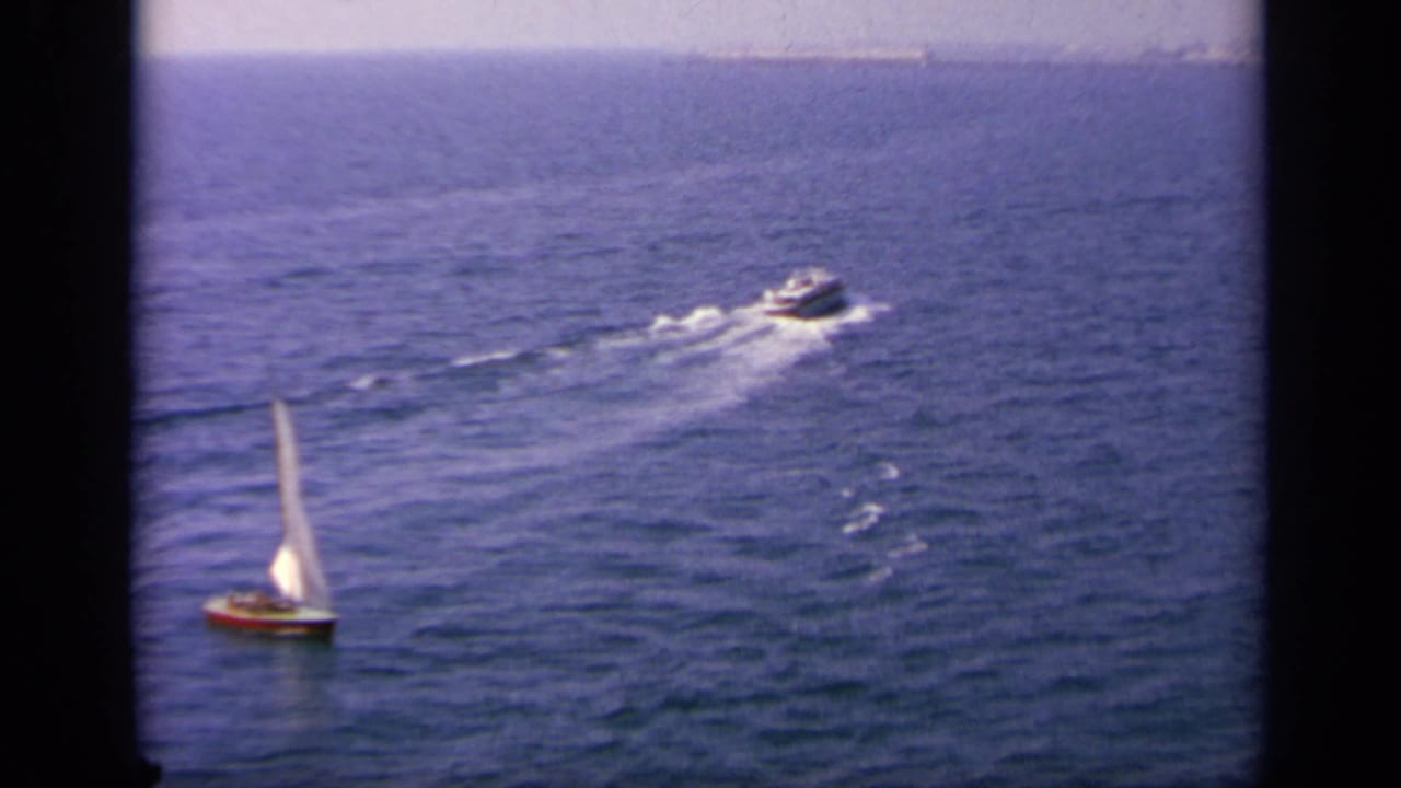 Elevated 1963 Greece-area 8mm view of a sailboat and a motorboat crossing open water, with wakes visible behind the moving boat.